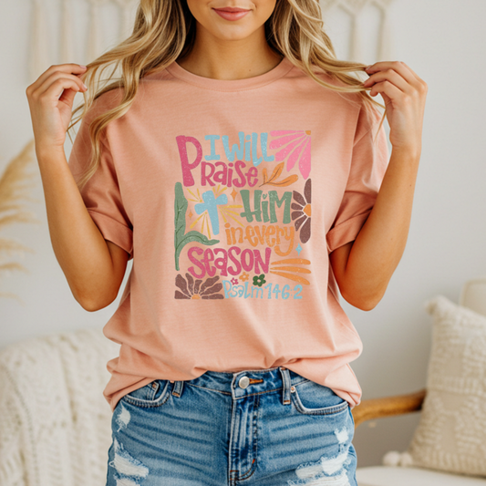 Person wearing a peach t-shirt with colorful text and design with text that says "I will praise him in every season"