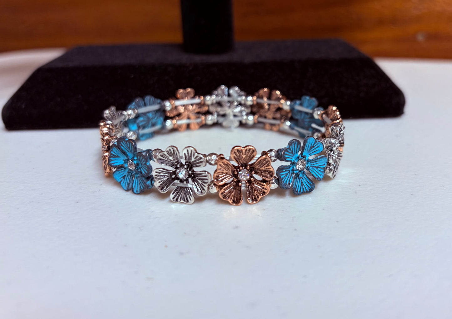 A close-up photograph of a floral stretch bracelet featuring alternating links of azure blue, silver, and copper-colored flowers. Each flower has a small clear crystal at its center. The bracelet is displayed on a white surface in front of a black velvet jewelry stand against a warm wood background.