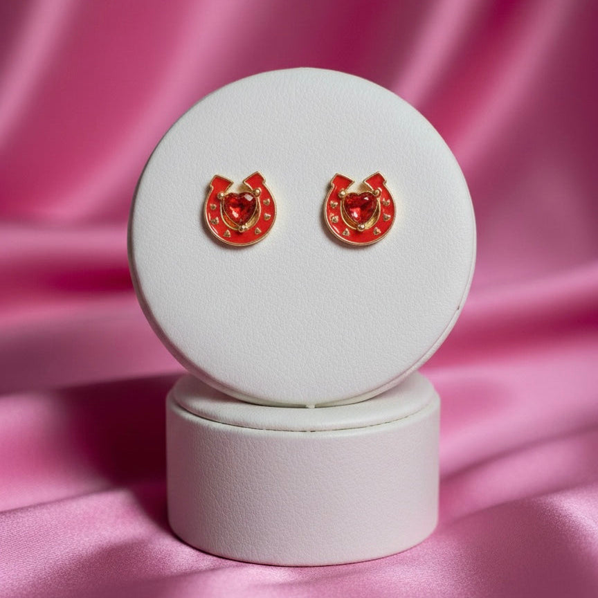 A pair of gold-toned horseshoe stud earrings displayed on a white circular stand. The horseshoes are filled with vibrant red enamel and feature a shimmering, heart-shaped red gemstone at the center. The earrings are set against a background of soft, draped pink satin fabric with elegant lighting.