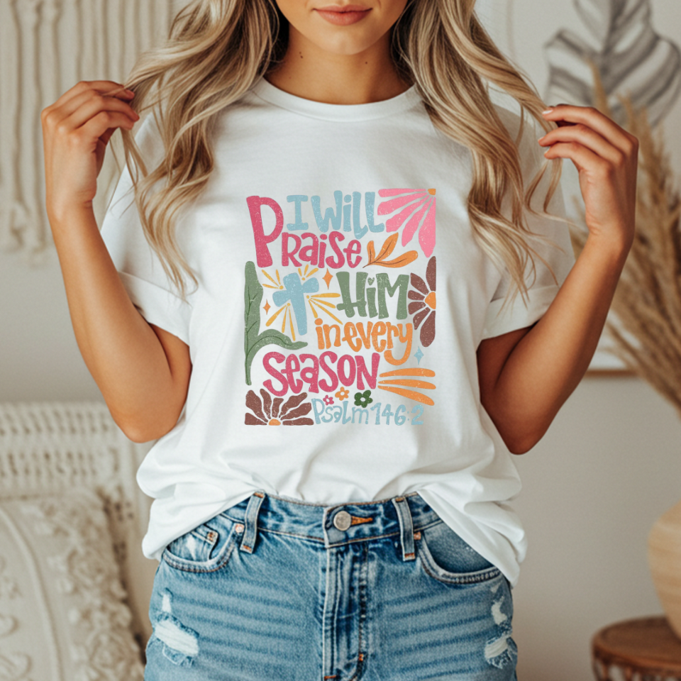 Person wearing a white t-shirt with colorful text and design with text that says "I will praise him in every season"