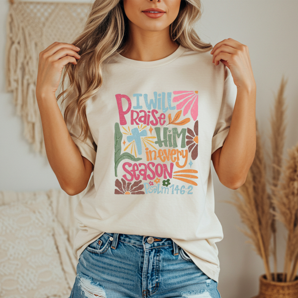 Person wearing a ivory t-shirt with colorful text and design with text that says "I will praise him in every season"