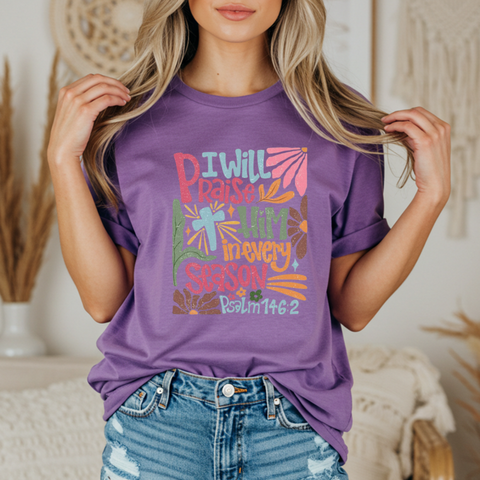 Person wearing a purple t-shirt with colorful text and design with text that says "I will praise him in every season"