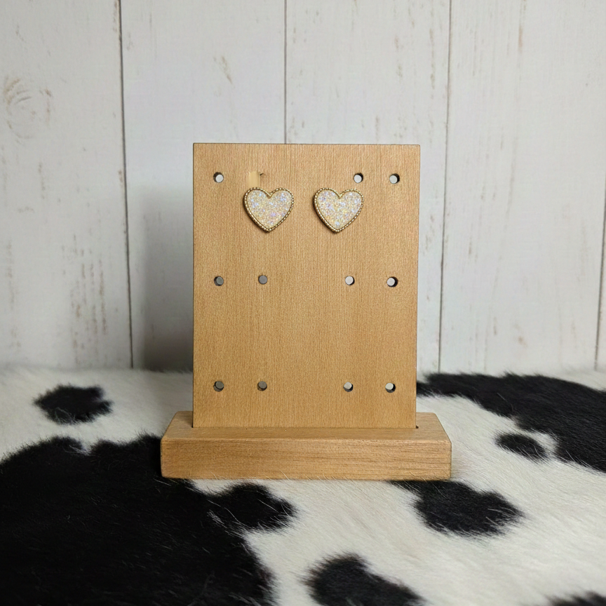 A pair of heart-shaped stud earrings featuring a shimmering champagne-gold glitter texture. The earrings are bordered with a thin, beaded-style gold metal rim and are displayed on a light wooden jewelry stand. The background includes vertical white wooden planks and a black-and-white cowhide patterned surface