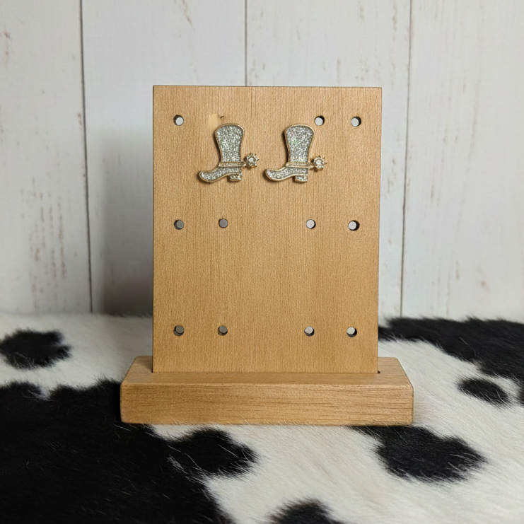 A pair of gold-toned cowboy boot stud earrings featuring a silver glitter-textured inlay. Each boot includes a small, gold star-shaped spur on the heel. The earrings are displayed on a light-colored wooden jewelry stand, which is set against a background of white wood paneling and a black-and-white cowhide patterned surface.