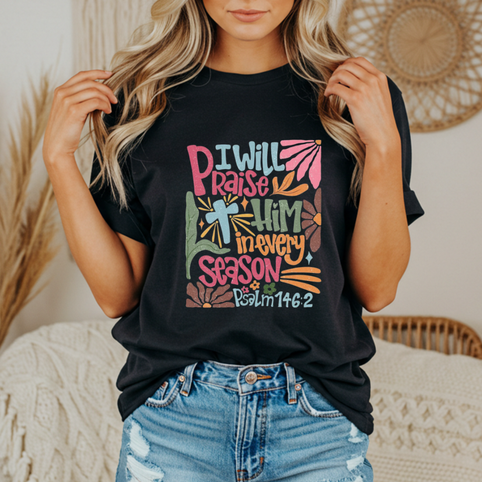 Person wearing a black t-shirt with colorful text and design with text that says "I will praise him in every season"
