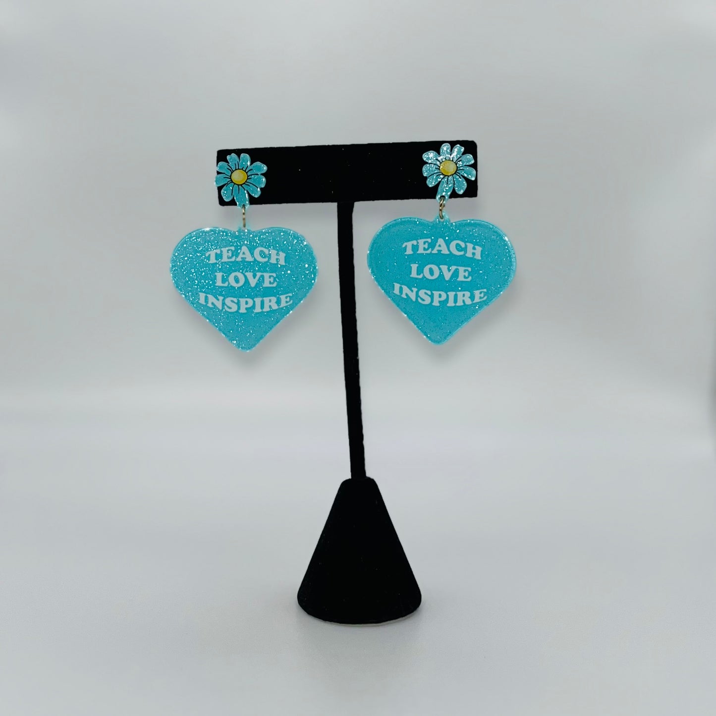 Heart-shaped earrings with 'Teach Love Inspire' text on a black stand against a white background