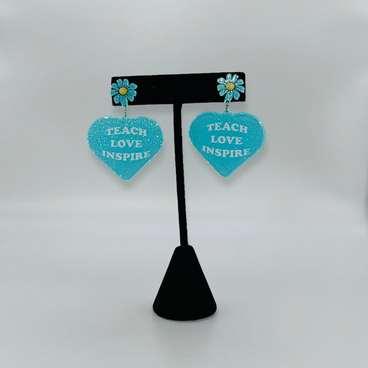 Heart-shaped earrings with 'Teach Love Inspire' text on a black stand against a white background