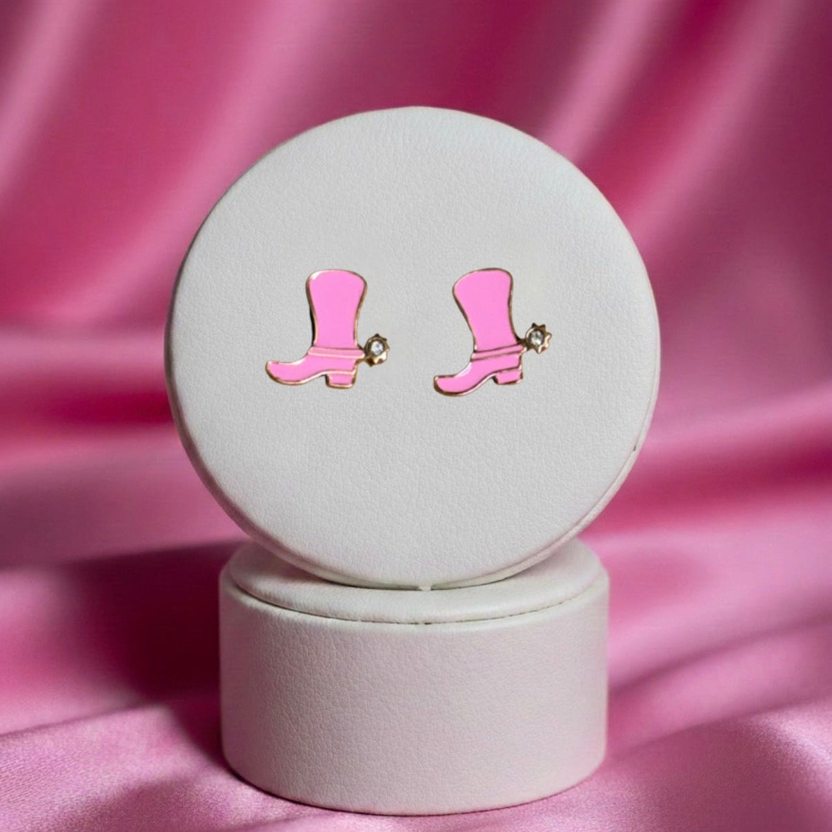 A pair of small, gold-toned stud earrings shaped like cowgirl boots with bright pink enamel filling. A tiny clear rhinestone is set into the heel of each boot to represent a spur. The earrings are displayed on a white circular jewelry stand against a draped, shimmering pink satin background.