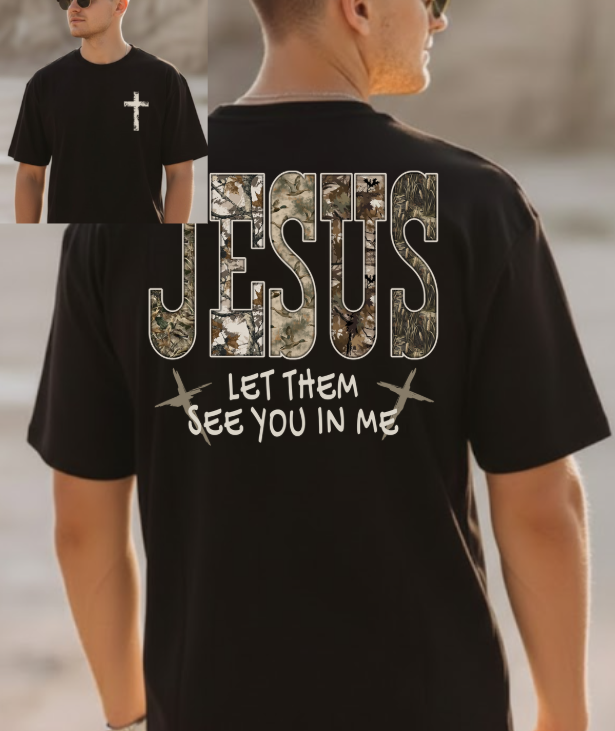 A man wearing a black short-sleeved t-shirt. The back of the shirt features the word "JESUS" in large, camo-patterned block letters, with the phrase "LET THEM SEE YOU IN ME" printed underneath in white, stylized font, flanked by two small crosses. An inset image shows the front of the shirt, which has a small, distressed white cross on the left chest.