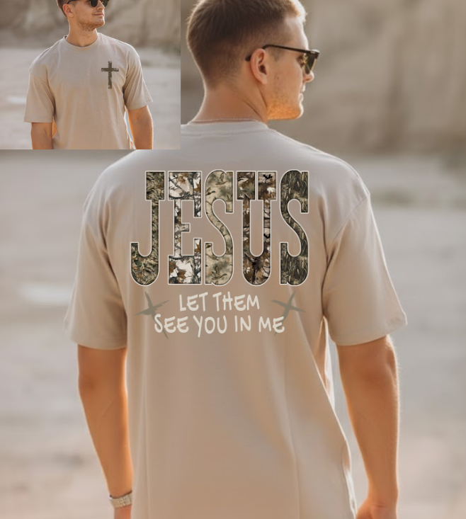 A man wearing a sand colored short-sleeved t-shirt. The back of the shirt features the word "JESUS" in large, camo-patterned block letters, with the phrase "LET THEM SEE YOU IN ME" printed underneath in white, stylized font, flanked by two small crosses. An inset image shows the front of the shirt, which has a small, distressed white cross on the left chest..