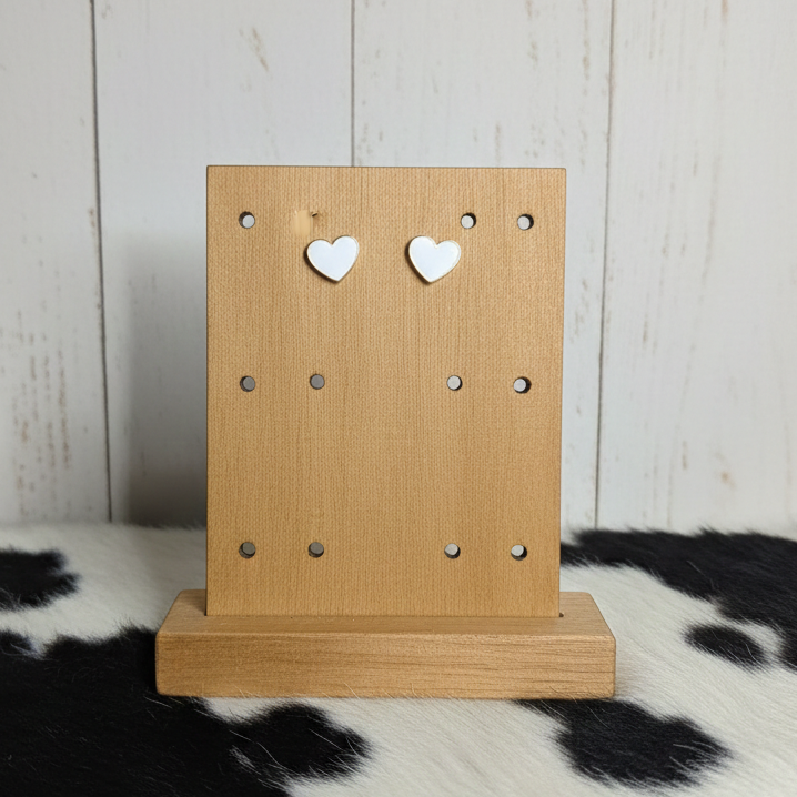 A pair of small, minimalist white heart-shaped stud earrings displayed on a light-colored wooden jewelry stand. The earrings have a smooth, matte white finish. The stand is placed against a background of vertical white wood paneling and a black-and-white cowhide patterned rug.